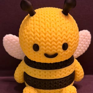 May include: A small, three-dimensional bee figurine with a textured, knit-like surface. The bee has a yellow body with two horizontal black stripes, black antennae, black eyes, a black smile, and two white wings extending from its sides.