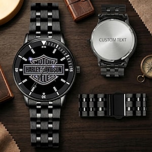 Op de afbeelding: Een zwart roestvrijstalen polshorloge met het Harley-Davidson-logo op de wijzerplaat, een bijpassende schakelarmband en een losse horlogekastbodem met de tekst CUSTOM TEXT gegraveerd op het ronde, zilverkleurige metalen oppervlak.