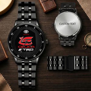 Könnte beinhalten: Eine schwarze Armbanduhr aus Edelstahl mit Gliederarmband, schwarzem Zifferblatt, Toyota-Logo, rotem Celica-Emblem und TRD-Schriftzug. Ein separates Uhrengehäuse zeigt den Text CUSTOM TEXT, zudem ist ein abgenommenes Uhrenarmband zu sehen.