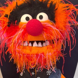 May include: A plush puppet character with bright orange fur, a large red nose, and wild orange and pink hair. The puppet wears a black bandana with a white paisley pattern, a blue denim vest, and a white rope belt. It holds a wooden drumstick.