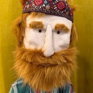 May include: A puppet with a light-colored felt face, small black eyes, and thick ginger faux fur hair and beard. The puppet wears a dark headband featuring a colorful floral pattern and a green, white, and red plaid button-down shirt.