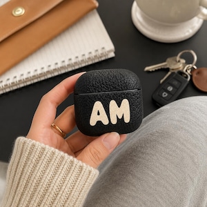 May include: A black, textured, square-shaped protective case for wireless earbuds. The front of the case features the letters AM in a large, cream-colored, rounded font. The case has a smooth, rounded edge design and a matte finish.