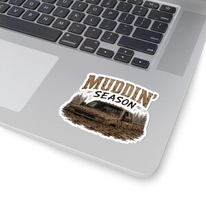 May include: A die-cut sticker featuring a pickup truck covered in brown mud, set against a forest background. The text MUDDIN' SEASON is printed in a textured, mud-like font above the truck. The sticker is placed on a silver laptop surface.