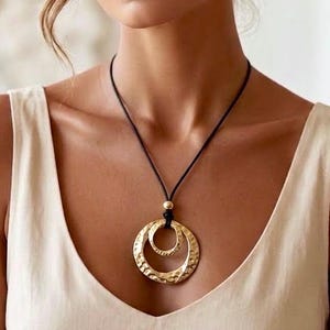 May include: A necklace featuring two interlocking gold-toned circular pendants with a hammered, textured finish. The pendants are suspended from a thin black cord, which is accented by a single small gold-toned bead positioned above the rings.