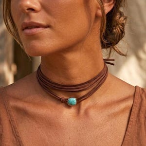May include: A brown suede cord choker necklace featuring a single square-shaped turquoise stone set in a silver-toned metal frame. The cord wraps multiple times around the neck and is secured with a small knot at the front near the stone.