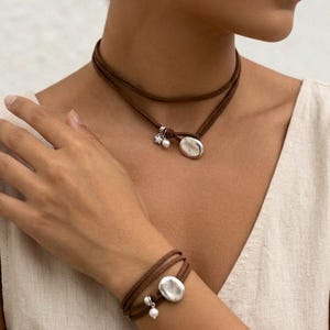 May include: A brown suede cord choker and matching wrap bracelet set. Both pieces feature a silver-toned oval charm, a small white pearl bead, and a tiny silver star charm. The jewelry is designed with multiple wraps around the neck and wrist.