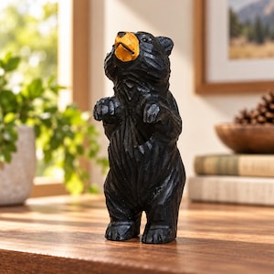 Peut inclure: Une figurine d'ours en bois sculptée à la main, debout sur ses pattes arrière avec les pattes avant levées. L'ours est peint en noir avec un museau jaune-orangé distinct, présentant des marques de ciseau visibles sur toute sa surface.