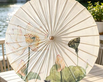 May include: A circular parasol made of off-white fabric featuring a hand-painted design of pink and white lotus flowers, green lily pads, and a dark seed pod. The umbrella has a wooden central hub and radiating ribs that create a pleated texture.