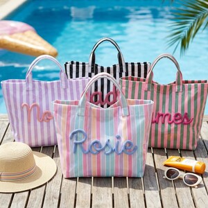 Peut inclure: Quatre sacs fourre-tout à rayures verticales de différentes couleurs, chacun portant un nom en lettres cursives en relief. Un sac est lavande, un noir et blanc, un arc-en-ciel pastel et un vert et rose. Un chapeau de paille, des lunettes et crème solaire.
