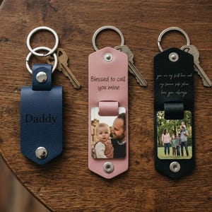 May include: Three leather keychains in blue, pink, and black with silver metal rings and keys. The blue one is embossed with the word Daddy. The pink one features the text Blessed to call you mine and a photo of a person holding a baby. The black one has printed text and a family photo.