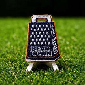 May include: A navy blue and orange embroidered patch shaped like a kitchen grater. The patch features a repeating pattern of white oval cutouts and the text BEAR DOWN in bold white letters. It is mounted on a small white plastic display stand.