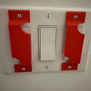 May include: Two bright orange plastic light switch guards mounted over the outer two switches of a triple rocker light switch plate. The guards are secured by the existing screws on the white wall plate, preventing accidental operation of the switches.
