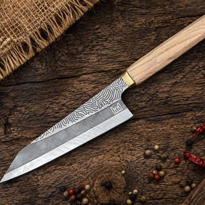 May include: A kitchen knife with a light-colored wooden handle and a brass bolster. The blade features a distinct dark gray finish with a complex, swirling white pattern along the spine and a sharp, straight cutting edge with a small logo.