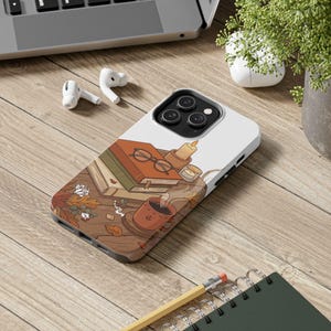May include: A white smartphone case featuring a printed illustration of a stack of books, a pair of round glasses, a steaming mug of coffee, and a lit candle, all arranged on a wooden surface decorated with scattered autumn leaves and berries.