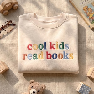 May include: A cream-colored crewneck sweatshirt featuring the embroidered text "cool kids read books" in multicolored letters. The sweatshirt is surrounded by a wooden toy car, brown-rimmed glasses, a storybook, wooden alphabet blocks, and a teddy bear.