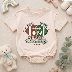 May include: A beige short-sleeved baby bodysuit featuring a graphic of a football split into brown and green halves. The text reads DAD MOM I'M STILL DECIDING. The design includes white laces, a logo, and three stars below the text.