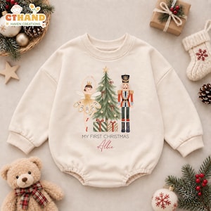 May include: A cream-colored long-sleeve baby romper featuring a watercolor graphic of a ballerina, a Christmas tree, and a nutcracker. Below the graphic, the text reads "MY FIRST CHRISTMAS" in black and the name "Albie" in red script.