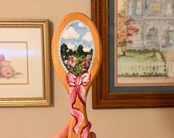 Escova de cabelo pintada à mão com desenho de paisagem e laço rosa.