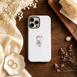 May include: A white smartphone case featuring a small, centered illustration of a grey and white tabby cat sitting and looking up at a tiny yellow butterfly. The case has a black camera lens cutout at the top left corner and a smooth finish.
