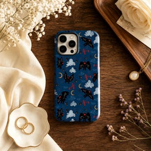 Könnte beinhalten: Eine dunkelblaue Smartphone-Hülle mit einem wiederkehrenden Muster aus schwarzen, mottenartigen Kreaturen mit roten Augen, hellblauen Wolken, kleinen gelben Mondsicheln und winzigen roten Flugzeugsilhouetten auf der Oberfläche.