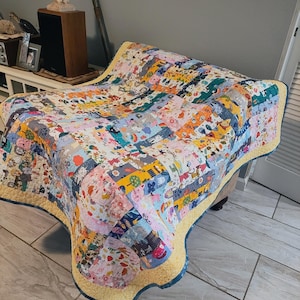 May include: A patchwork quilt featuring a variety of colorful fabric squares with diverse patterns including animals, rainbows, and floral motifs. The quilt is finished with a bright yellow border and a dark blue edge, showcasing intricate stitching.