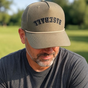 May include: An olive green baseball cap featuring the word BASEBALL embroidered in black capital letters across the front panel. The cap includes a decorative off-white cord running horizontally across the base of the crown above the brim.
