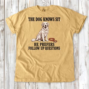 May include: A mustard yellow short-sleeve t-shirt featuring a graphic of a sitting golden retriever dog and a brown leash. The text reads: THE DOG KNOWS SIT. HE PREFERS FOLLOW UP QUESTIONS. The shirt has a crew neckline and a relaxed fit.