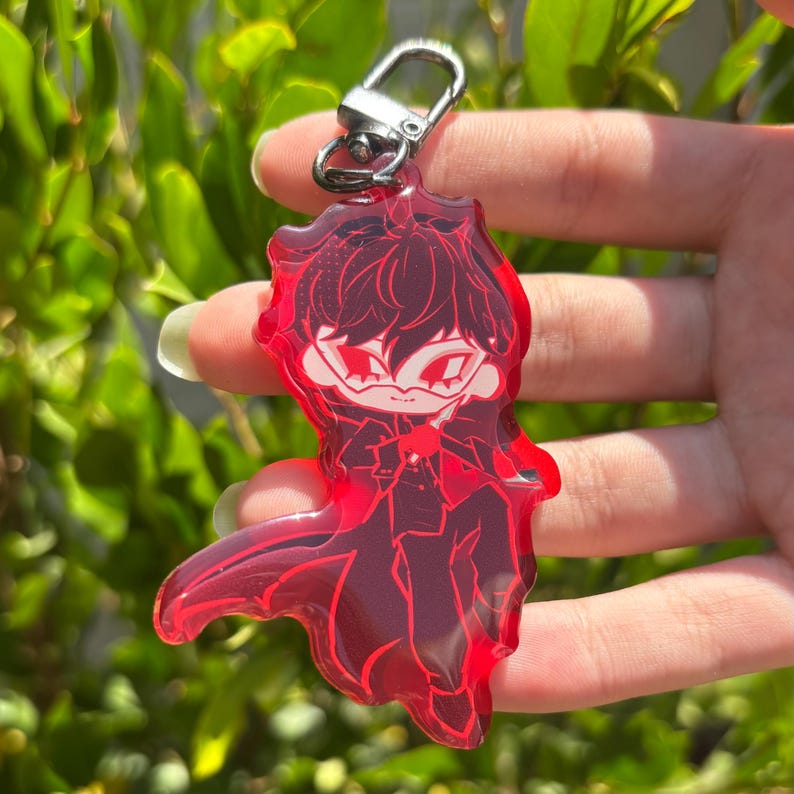 May include: A translucent red acrylic keyring featuring a stylised anime character with dark hair and a mask, wearing a long coat. The character is depicted in a dynamic pose with sharp lines and a vibrant red colour scheme, attached to a silver metal clasp.