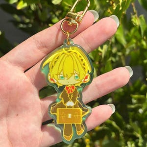 May include: A clear acrylic keyring featuring a stylised illustration of a character with bright yellow hair, wearing a dark brown outfit with a red bow tie, and holding a yellow rectangular briefcase, attached to a gold-toned metal lobster clasp.