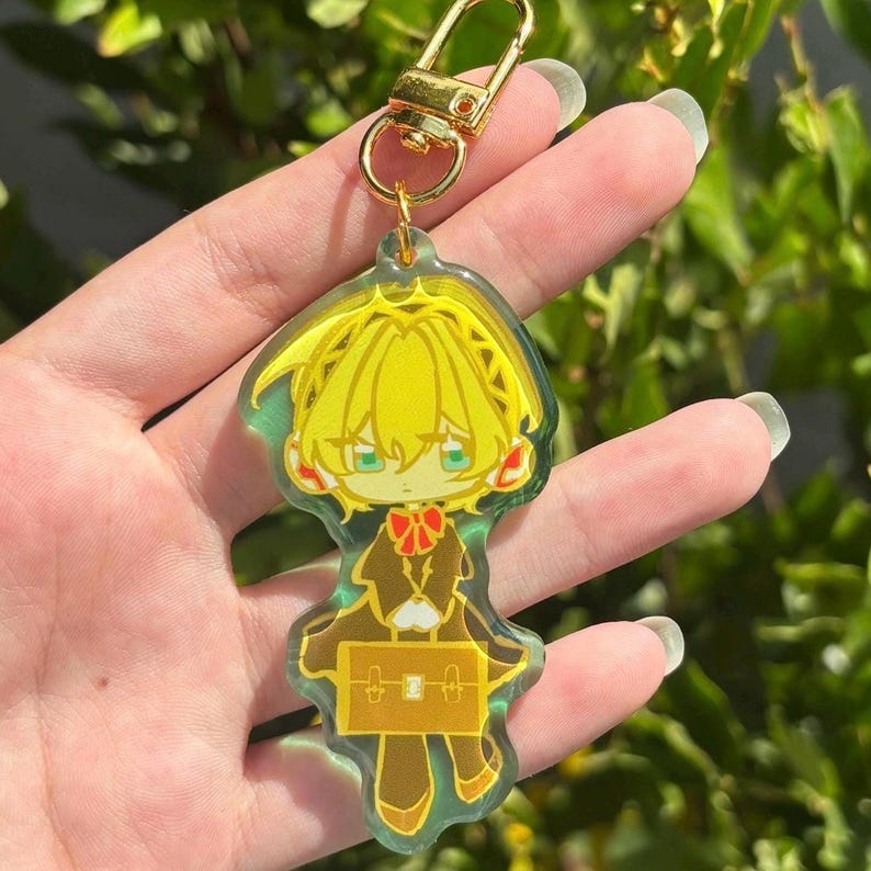 May include: A clear acrylic keyring featuring a stylised illustration of a character with bright yellow hair, wearing a dark brown outfit with a red bow tie, and holding a yellow rectangular briefcase, attached to a gold-toned metal lobster clasp.