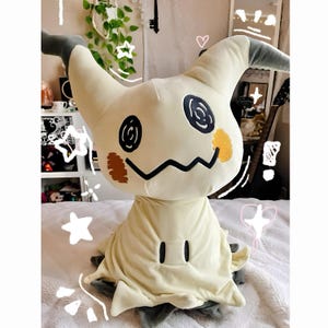 May include: A plush toy resembling a ghost-like creature with a pale yellow, fabric-covered body, two pointed ears with dark gray tips, and two black circular eyes. It features a jagged black mouth, two small black rectangular markings, and orange patches.