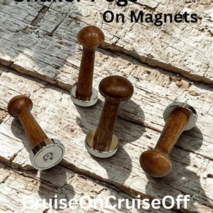 May include: Four brown wooden Shaker pegs with silver-colored circular magnetic bases. The text Shaker Pegs On Magnets and CruiseOnCruiseOff is visible. Each peg features a rounded top and a cylindrical stem attached to a flat metal magnet.