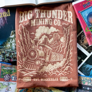May include: A rust-coloured t-shirt featuring a cream-coloured graphic of a steam locomotive train travelling through a canyon. The text reads BIG THUNDER MINING CO. and EST. MDCCCXLIX, with two sticks of dynamite flanking the bottom date.