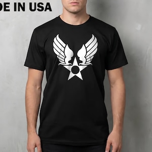 May include: A black short-sleeved t-shirt featuring a white graphic of a star with wings and a central circle. The text MADE IN USA is printed in the upper left corner. The shirt has a crew neckline and a standard fit.