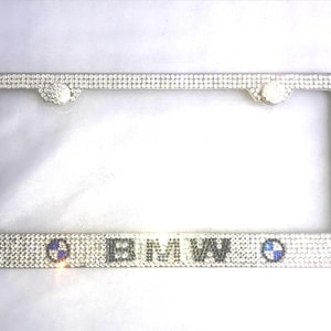 May include: A rectangular license plate frame covered in clear, reflective rhinestones. The frame features the text BMW in black lettering centered at the bottom, flanked by two circular blue and white emblems on either side of the letters.