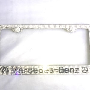 May include: A rectangular license plate frame covered in clear, reflective rhinestones. The bottom section features the text Mercedes-Benz in a dark font, flanked by two circular logos, all set against a bright, shimmering, crystal-encrusted surface.