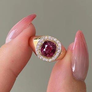 Puede incluir: Un anillo de oro con una piedra preciosa central de color rosa intenso en talla cojín, rodeada por un halo de pequeñas piedras claras en talla redonda. La banda es lisa y pulida, mostrando un diseño clásico con una piedra central vibrante.