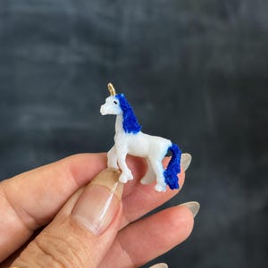May include: A small white unicorn figurine featuring a vibrant blue mane and tail with a textured, fuzzy appearance. The unicorn has a single, pointed gold-colored horn on its forehead and is depicted in a standing position with four legs.