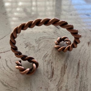 Op de afbeelding: Een koperkleurige metalen armband met een doorlopend gedraaid touwontwerp. De armband heeft een open structuur waarbij beide uiteinden naar binnen krullen in kleine, strakke spiralen, wat een opvallend S-vormig silhouet creëert.