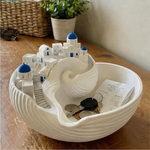 Peut inclure: Un bol décoratif blanc présentant une scène de village méditerranéen sculptée avec des bâtiments à dômes bleus et des escaliers en colimaçon intégrés dans une structure en forme de coquillage. Le bol contient des clés et un reçu en papier.