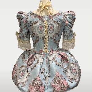 May include: A light blue historical dress with a floral pattern in pink and brown. The dress features short, puffed sleeves, a laced back, and a full skirt. The neckline is adorned with a yellow bow, and the sleeves have decorative trim.