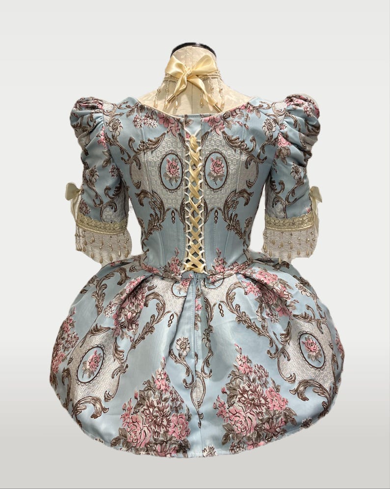 May include: A light blue historical dress with a floral pattern in pink and brown. The dress features short, puffed sleeves, a laced back, and a full skirt. The neckline is adorned with a yellow bow, and the sleeves have decorative trim.