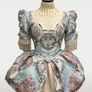 May include: A light blue historical dress with a floral pattern in pink and brown. The dress features puffed sleeves, a fitted bodice, and a full skirt. The dress is displayed on a mannequin.