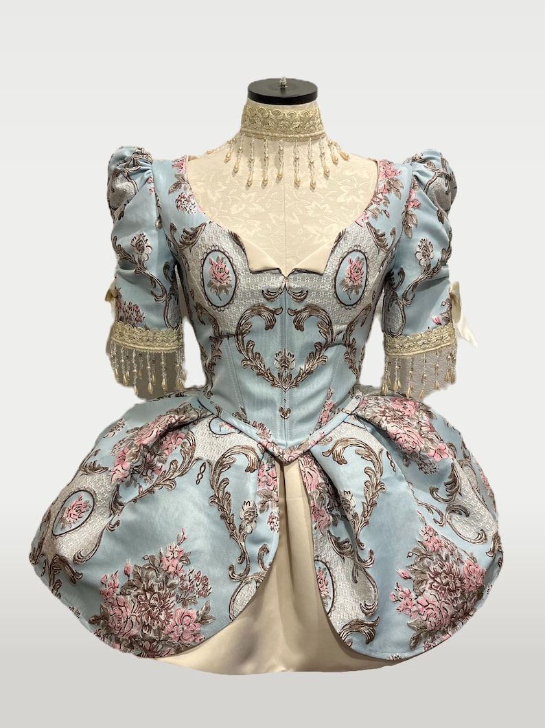 May include: A light blue historical dress with a floral pattern in pink and brown. The dress features puffed sleeves, a fitted bodice, and a full skirt. The dress is displayed on a mannequin.
