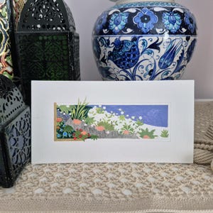 May include: A rectangular white card featuring a horizontal illustration of a garden scene with green foliage, small pink and blue flowers, and a solid blue sky section. The artwork is framed by a thin gold border on the left and bottom edges.