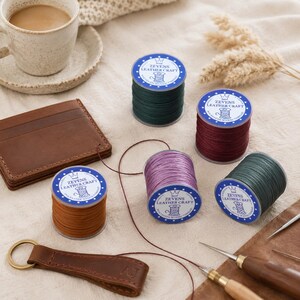 May include: Five spools of Zevens Leather Craft polyester thread in dark green, burgundy, brown, and purple, arranged with a brown leather card holder, a leather keychain, and two metal awls. Each spool features a blue label with white text.