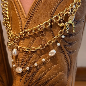 May include: A gold-toned metal chain boot accessory featuring a horseshoe charm with the text GOOD LUCK, dangling white faux pearls, and small metallic links. The accessory is draped over a tan leather western-style boot with decorative stitching.