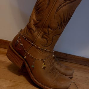 Könnte beinhalten: Ein beigefarbener Cowboystiefel aus Leder mit aufwendigen Ziernähten, verziert mit einer mehrlagigen, goldfarbenen Ketten-Fußkette. Die Fußkette ist mit kleinen Anhängern versehen, darunter ein Miniaturhut, ein Kaktus und bunte Perlen.