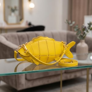 May include: A bright yellow fish-shaped crossbody bag with a segmented body design, a top zipper closure, and a matching yellow shoulder strap with gold-toned metal hardware. The bag features a distinct tail fin and side fins, with a structured form.