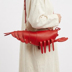 May include: A bright red shoulder bag shaped like a lobster, featuring segmented body panels connected by gold-toned metal rivets, a long adjustable shoulder strap, and dangling leg-like appendages extending from the bottom of the main compartment.